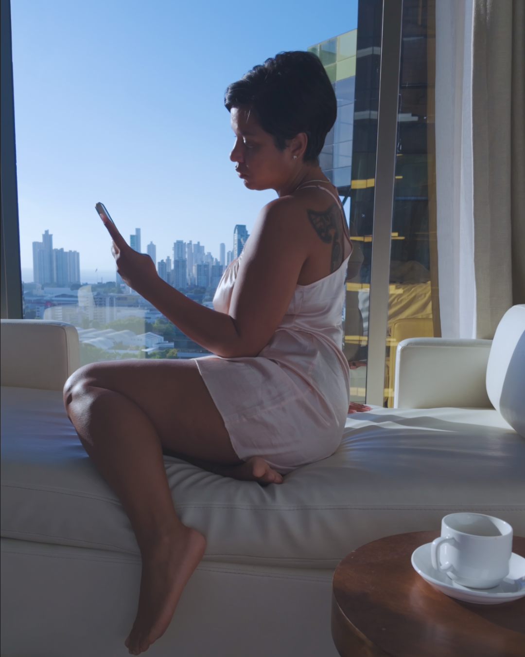 Woman in a white robe using a phone by a large window with a cityscape view.