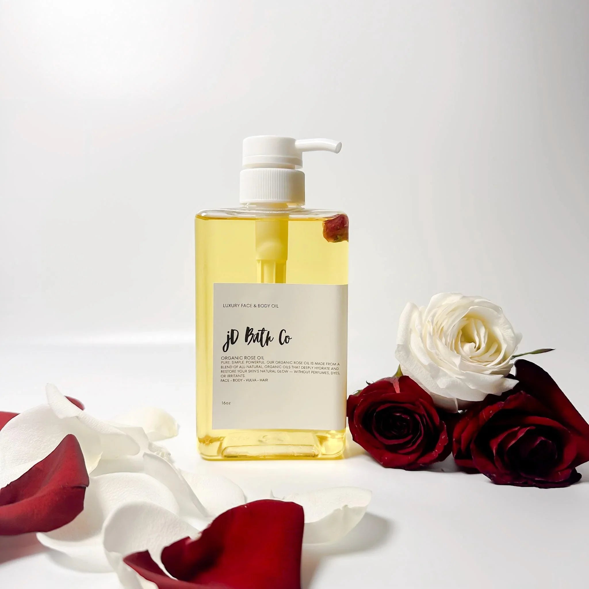 Bottle of jD Bath Co. rose body oil with real red rose buds. Red and white roses on a white background